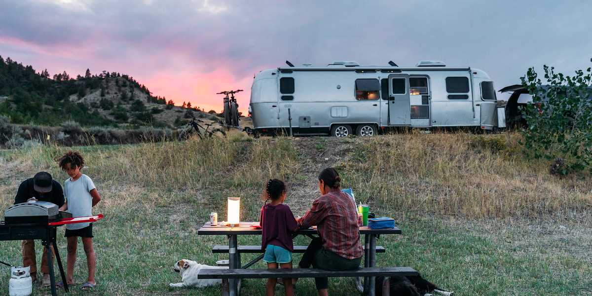 People relaxing at a picnic table on a grassy campsite near an RV at sunset in Montana
