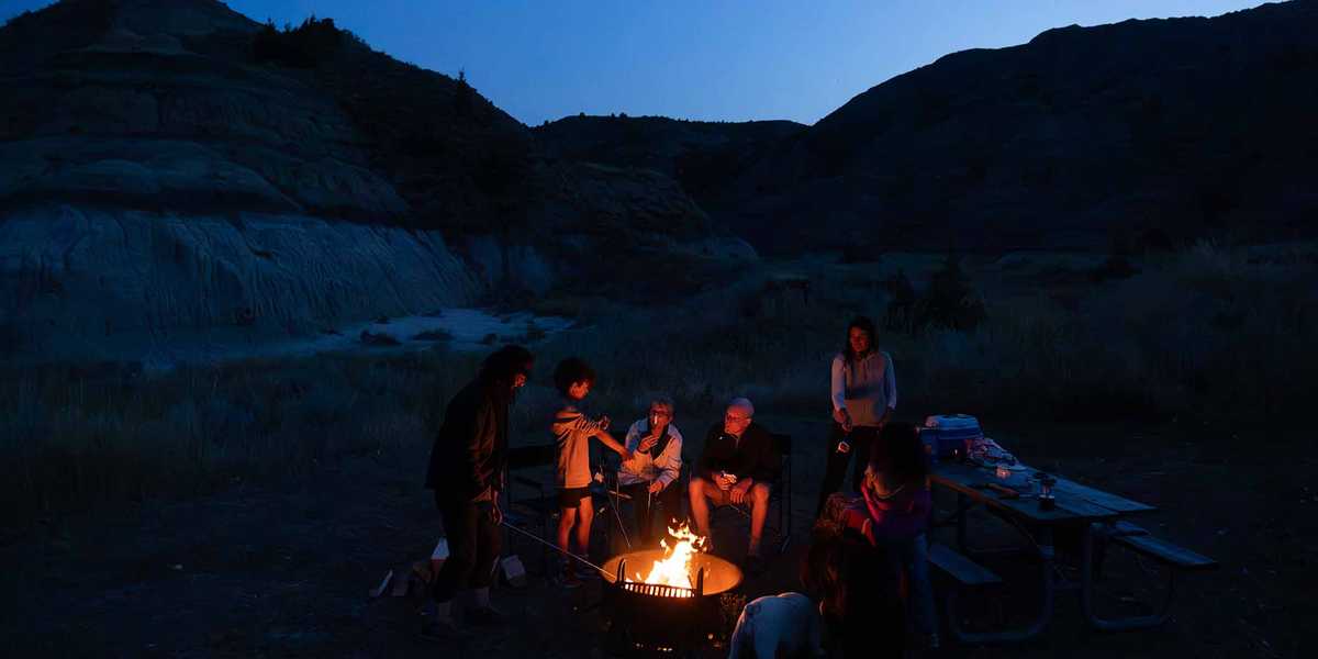 Group sitting around a campfire at dusk in a Montana canyon with mountains in the background