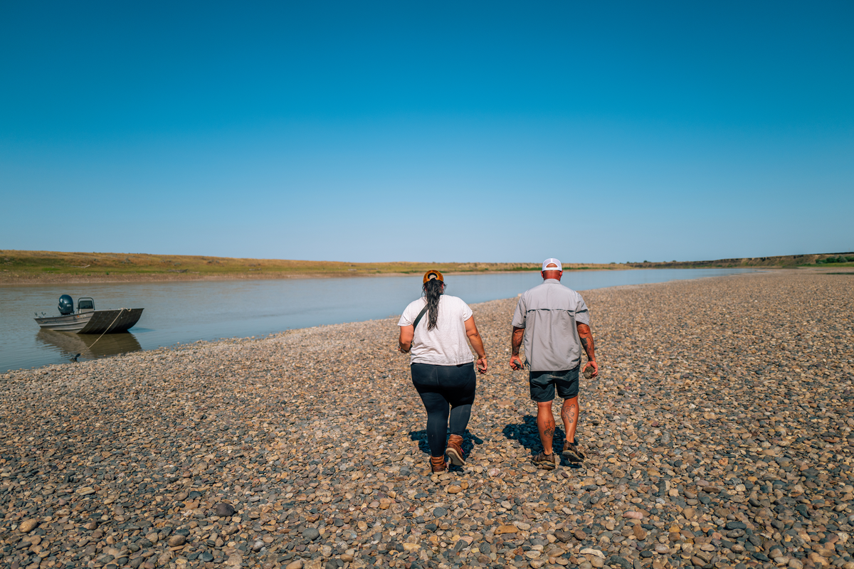 Two people walking along a rocky riverbank beside a small boat on a calm Montana river under a blue sky