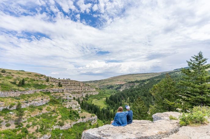 Person overlooks a green Montana valley from a rocky viewpoint under partly cloudy skies