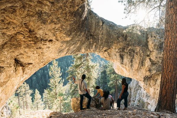 Hikers pause at a natural rock tunnel in a forested Montana canyon near a group of towering rock walls