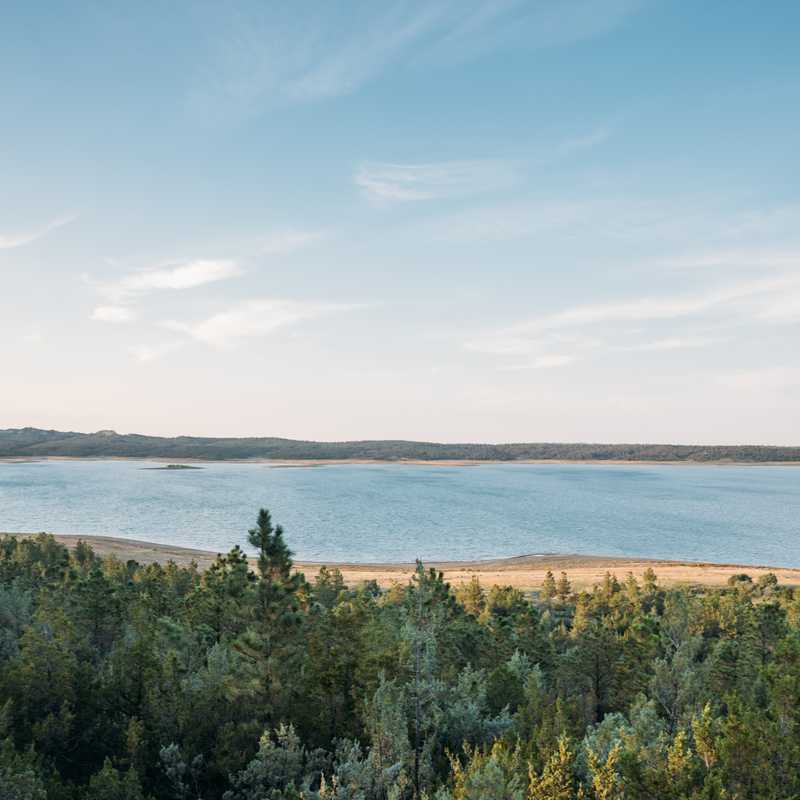 Wide view of a Montana river and shoreline bordered by forest and rolling hills under a blue sky