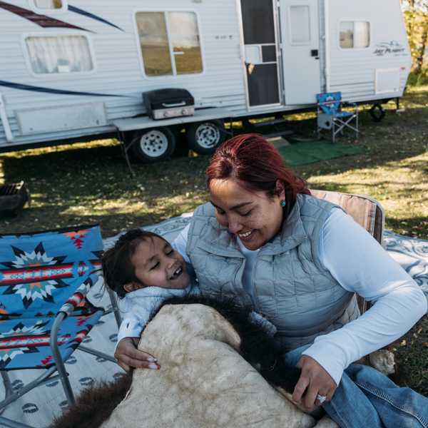 Two people relax outdoors at a campsite in Montana, one sitting on an outdoor chair and the other hugging while lying on a blanket near a trailer and picnic tables