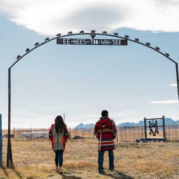 Two people walking through a metal archway at a rural ranch entrance in Montana, with mountains in the background and a sky-stamped slogan on the arch