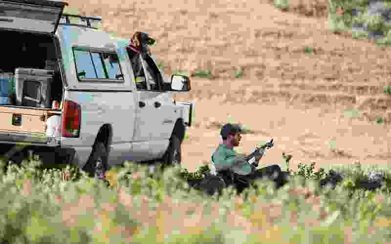 Two people camping by a pickup truck in a grassy Montana landscape, with one playing a guitar