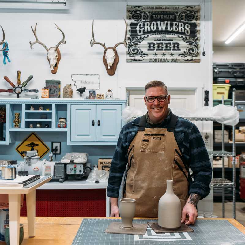 Man wearing an apron pours beer at a Montana brewing shop counter with “Growlers Beer” signage in the background