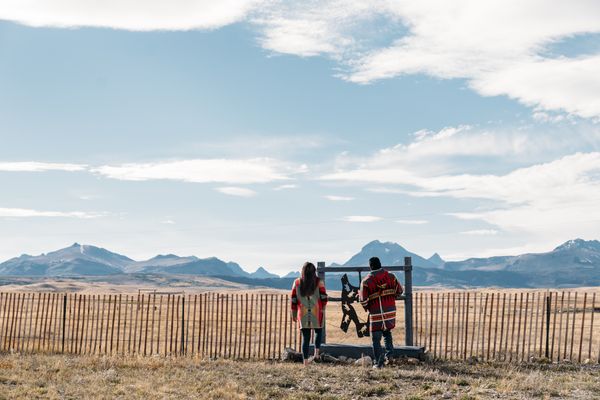 Two hikers walking along a fenced overlook with mountain peaks in the distance under a blue sky