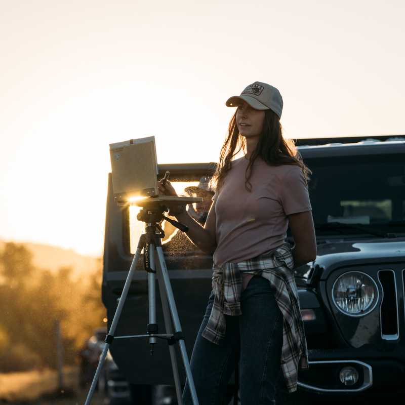 Woman standing near a Jeep with a camera tripod at sunset on a Montana road trip