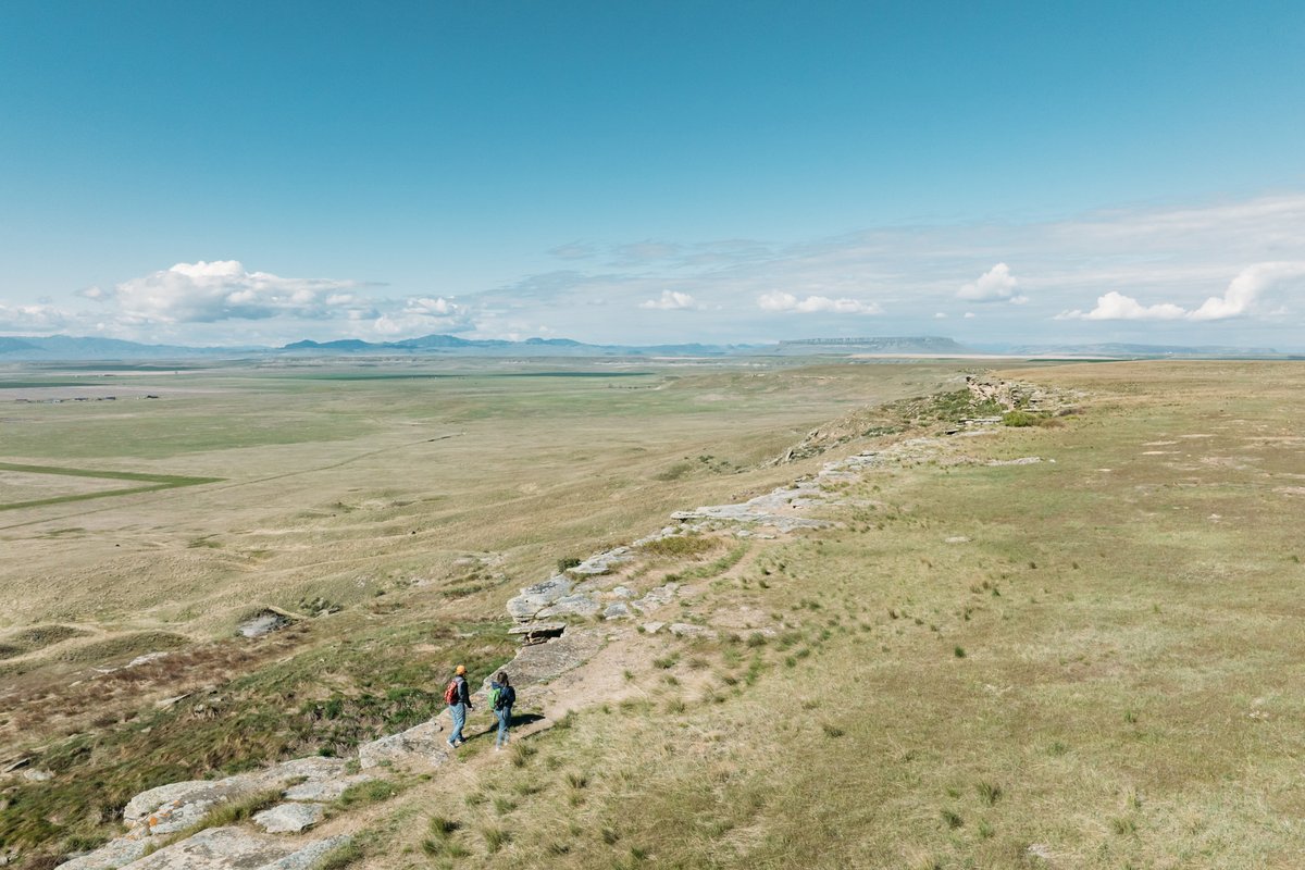 Hikers on a grassy ridge overlooking Montana plains and distant mountains under a blue sky