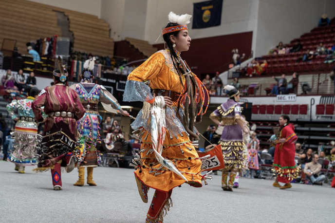 Native dancer in traditional regalia walking at an indoor powwow, with other dancers and spectators seated in the background