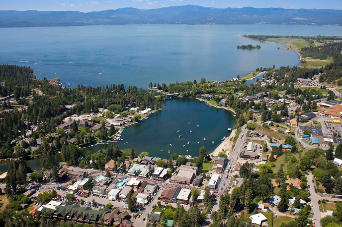 Aerial view of a Montana town along the shoreline of a large lake, with a marina and boats along the river inlet and surrounding forests