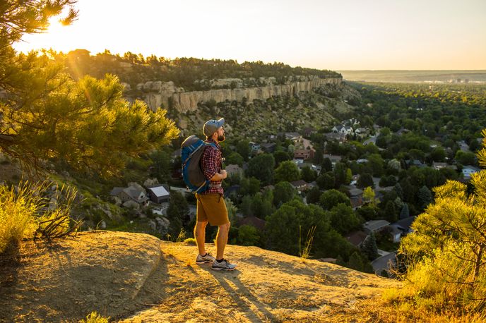 A person with a backpack stands on the rims, admiring a sprawling landscape of trees and houses at sunset.