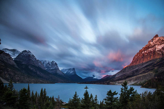 Lake and mountain peaks in western Montana at dusk, with pine trees along the shoreline under a colorful sky