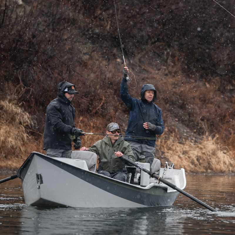 People fishing from a small boat on a Montana river with banks of dry shrubs and trees