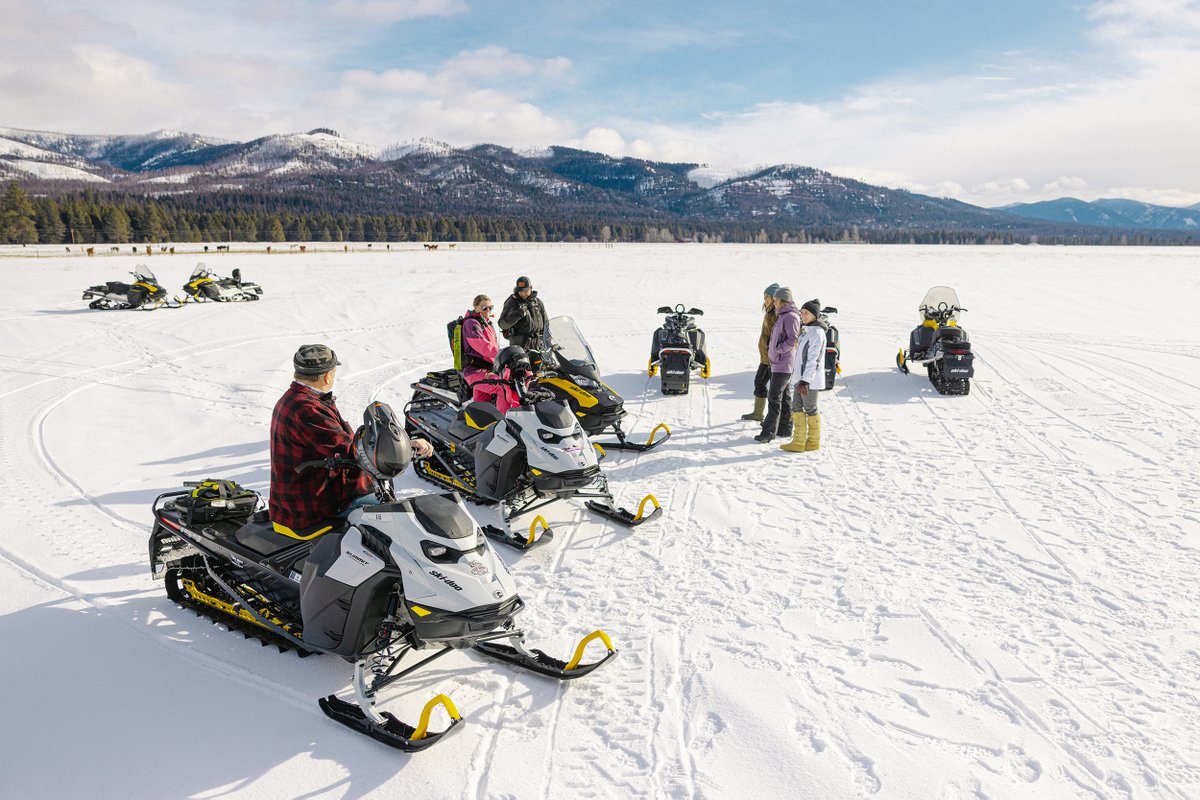 Snowmobiles parked on a snow-covered trail with riders along Montana’s Flathead Lake shoreline and mountains in the background