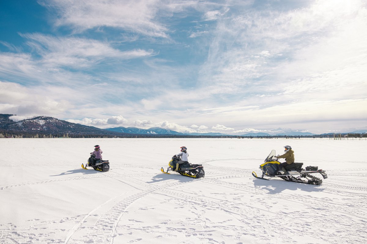 Snowmobiles on a snow-covered Montana lake with distant mountains under a partly cloudy sky