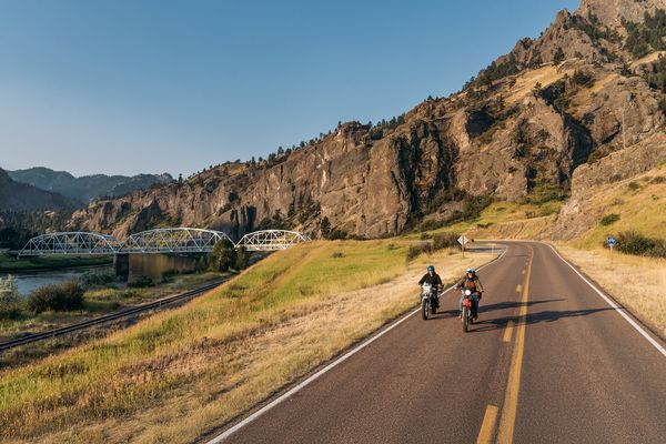 Motorcyclists ride along a mountain road beside the Missouri River, with a historic bridge in the background near Fort Benton, Montana