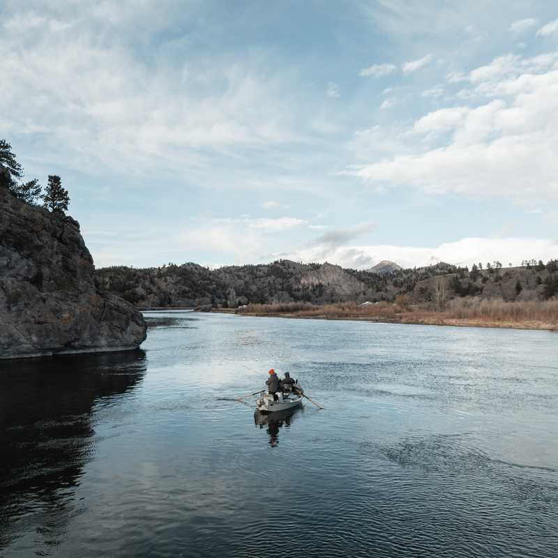 Boat on a calm river with rocky riverbanks and snow-dusted mountains in the distance under partly cloudy skies