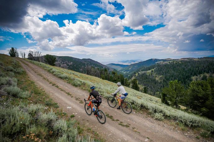 Cyclists ride a dirt road through Montana’s green hills and forests under dramatic clouds