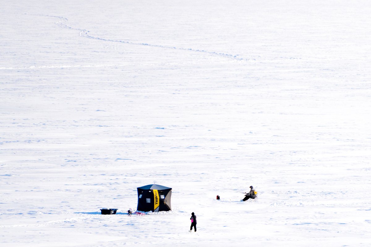 Snow-covered lake with a snowmobile near a shelter and two people ice skating in the distance, with mountain peaks and blue sky in the background