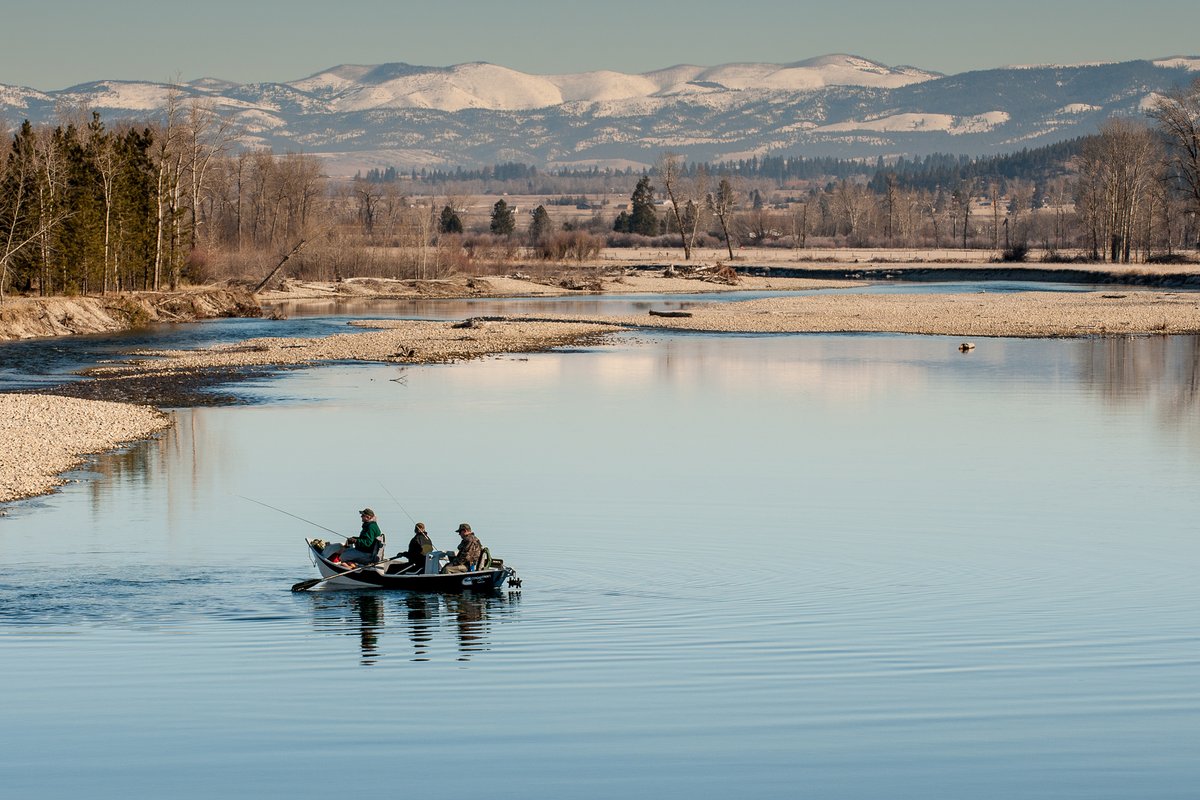 Boat with people on calm river waters near a Montana countryside shoreline with distant mountains in winter sunlight