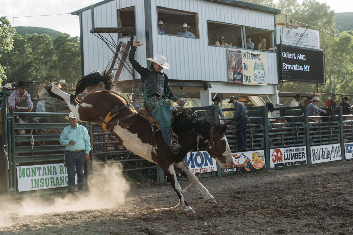Ranch rodeo action with a rider on a bucking horse in front of a barn-like arena and spectators, with Montana Ranch Insurance banners along the fence