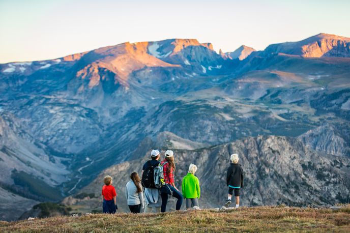 A group of people in colorful jackets admire a vast mountain landscape at sunset from a grassy viewpoint.
