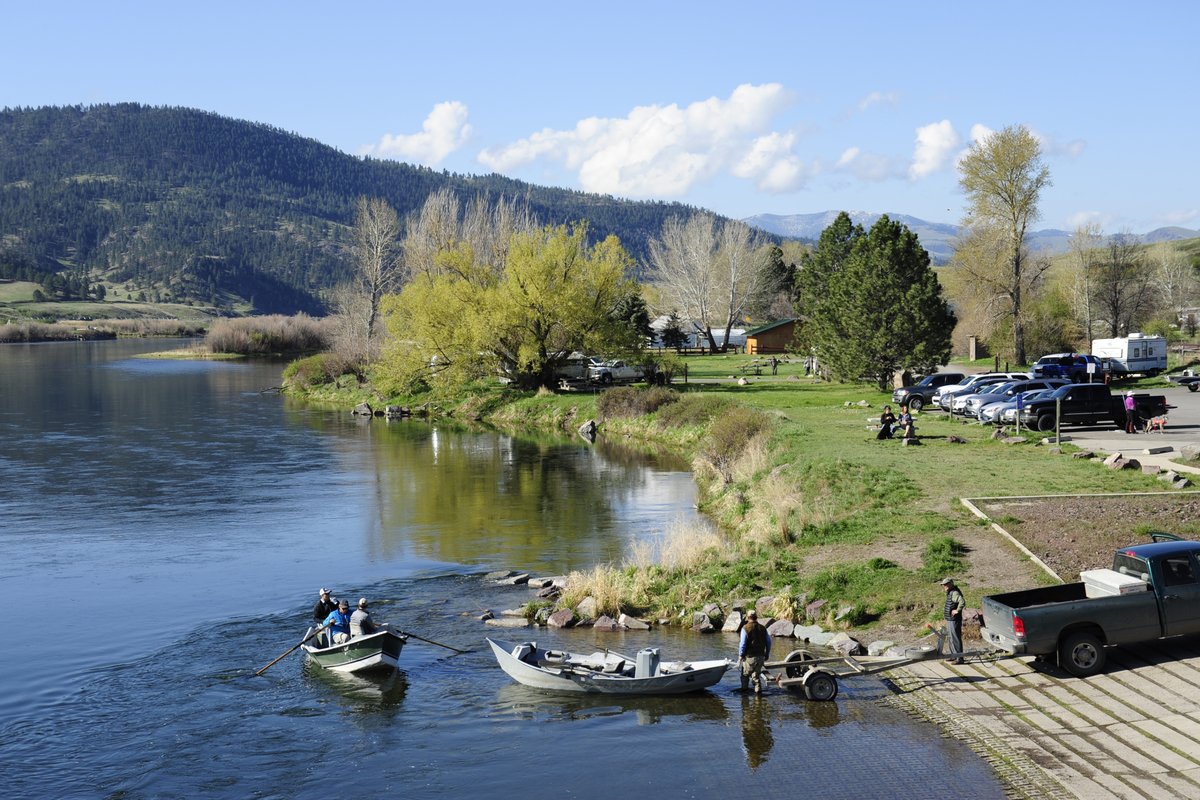 Boats and people by the shore along a calm river at a Montana lakeside campground, with mountains in the background