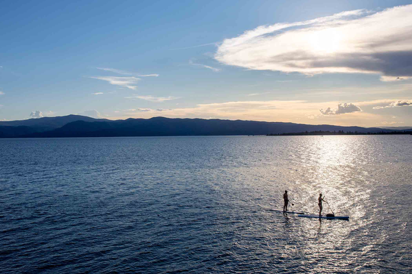 Two paddleboarders glide on calm water at a Montana lake near the shoreline at sunset, with distant hills and clouds in the sky
