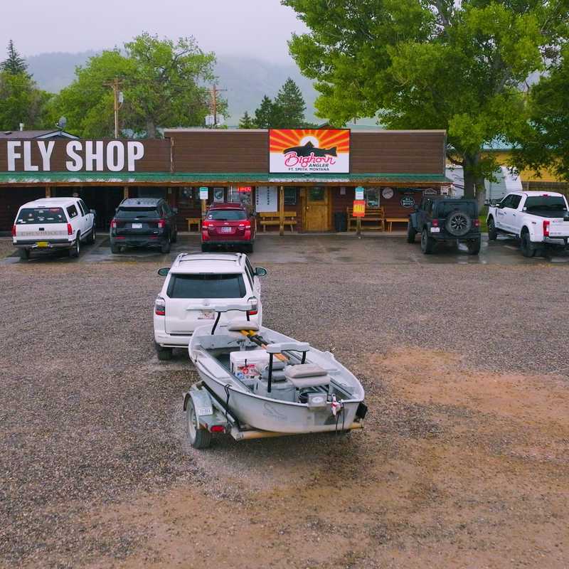 Parking lot in front of a Montana fly shop with boats on trailers and vehicles lined up near the storefront