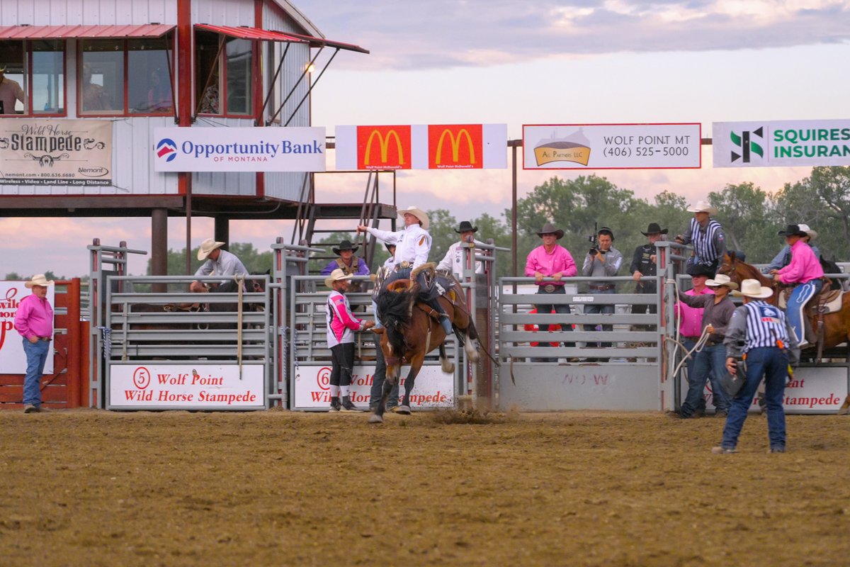 Riders and families at a rodeo arena with horses and spectators, backed by sponsor signs in Montana