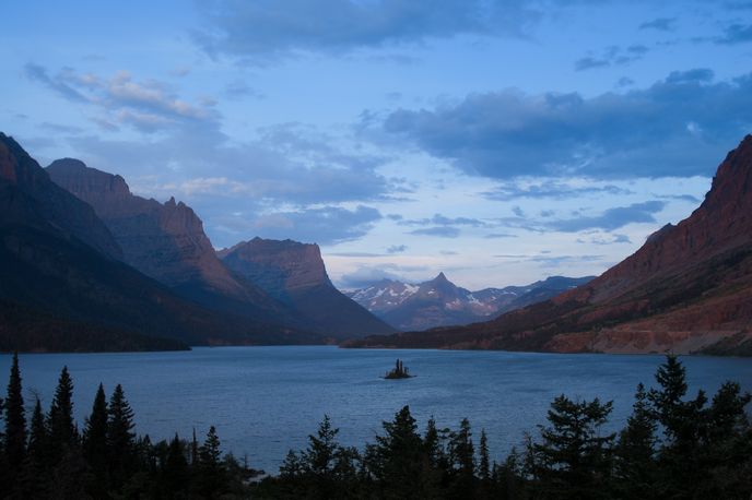 Scenic view of a calm mountain lake with red cliffs and snowcapped peaks at dusk in Montana