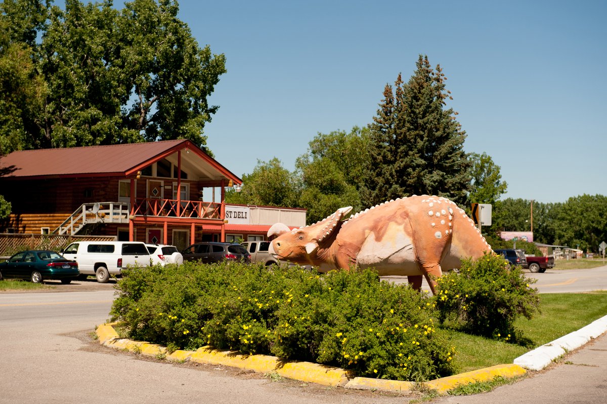 Giant dinosaur statue along a road outside a roadside building in Montana, with trees and parked vehicles nearby