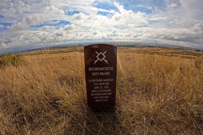 A gravestone in a grassy field with cloudy skies, commemorating Noisy Walking, a Cheyenne warrior who fell in 1876 defending his homeland.