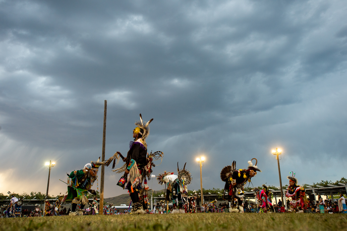 Native dancers in colorful regalia perform outdoors in a dusk sky at a Montana powwow
