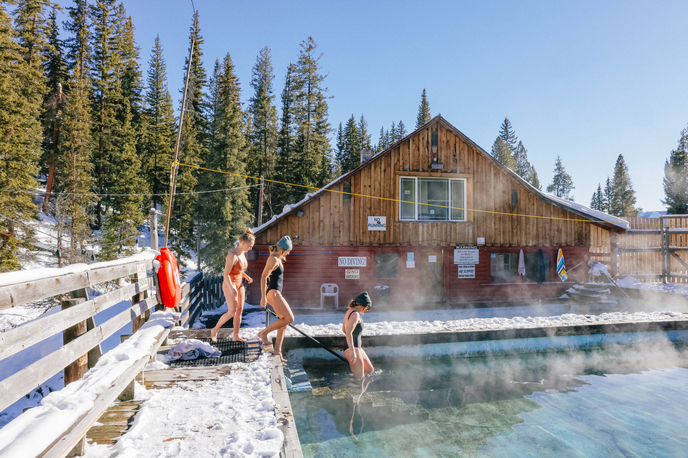 People relax in a snowy outdoor hot springs pool in front of a wooden bathhouse in Montana