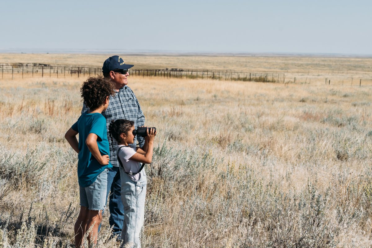 Two adults and a child stand together outdoors on Montana grasslands near a fence line under open skies