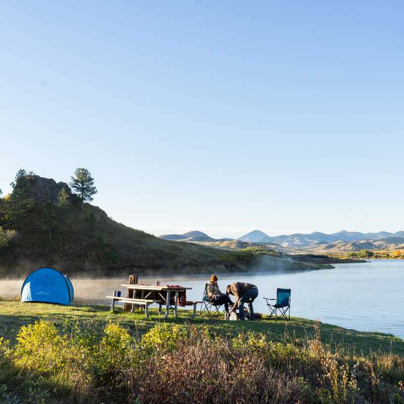 Camping along a calm Montana lake with a blue tent, picnic table, and people enjoying the shoreline at sunrise