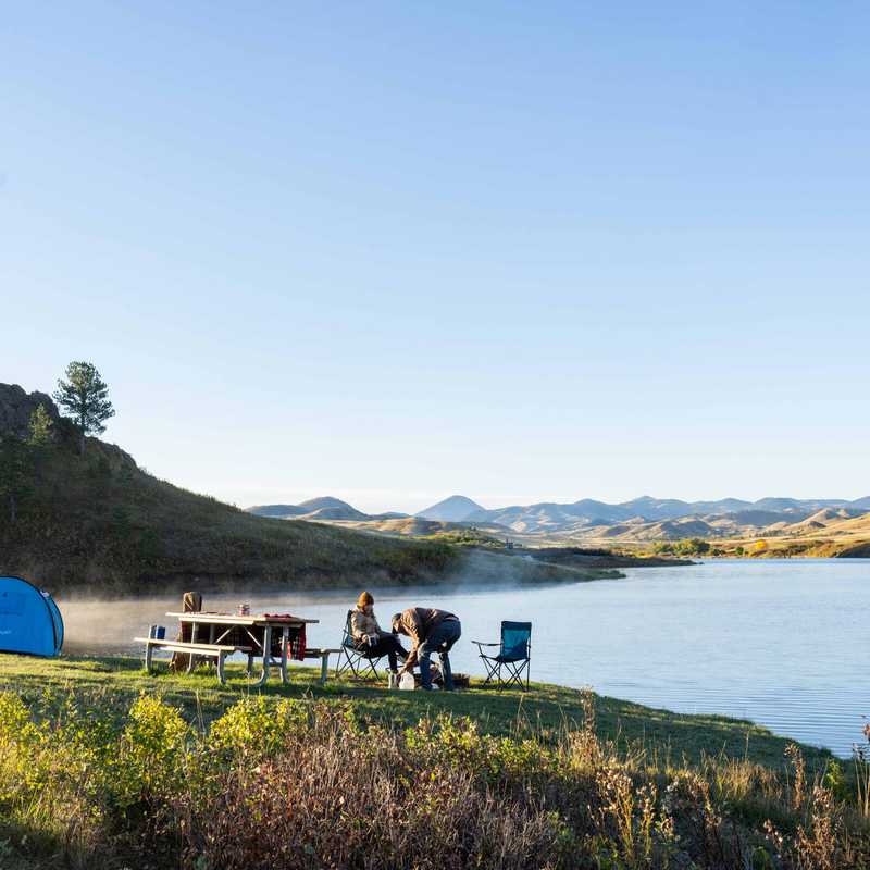 Camping setup with a blue tent and chairs along a calm lake shoreline, with mountains in the distance