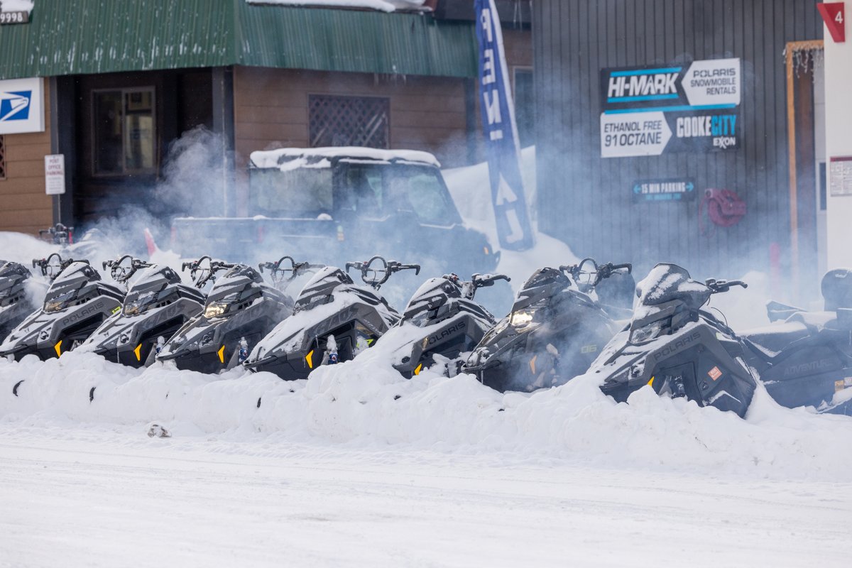 Row of snowmobiles parked outside a Montana building, with steam rising in the snowy parking area