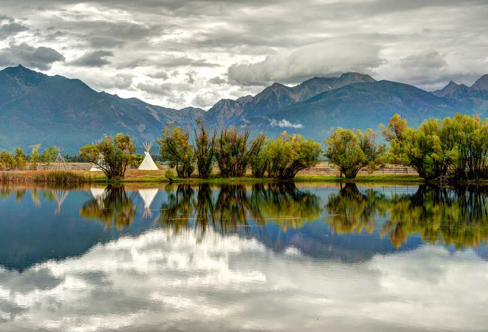 Calm lake reflecting mountains and a cloudy sky with a row of trees along the shore in Montana