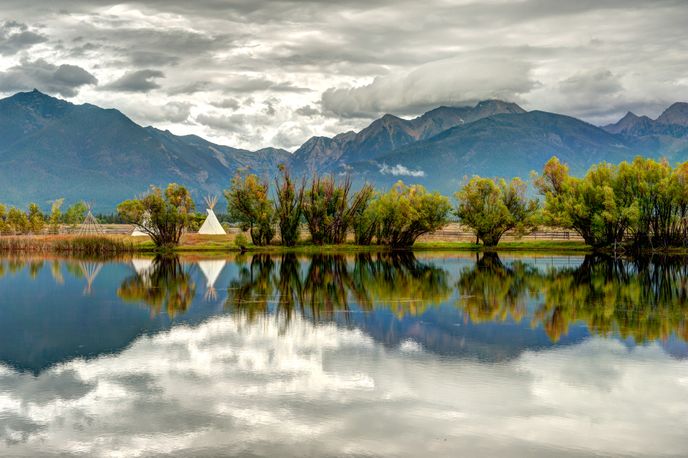 Calm lake reflecting mountains and a cloudy sky with a row of trees along the shore in Montana
