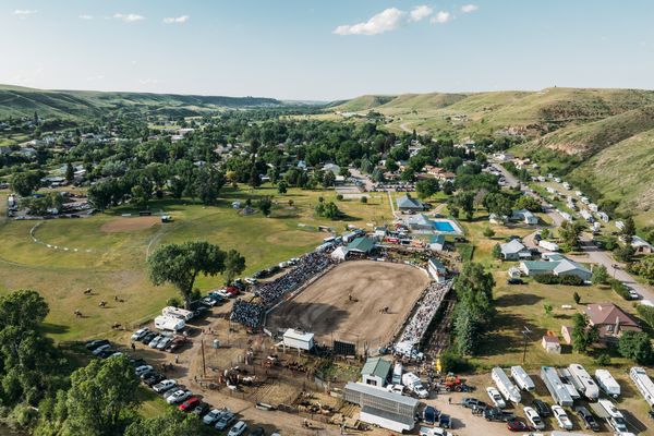 Aerial view of a small Montana town with a central event grounds and nearby RV parking along a main road amid rolling hills