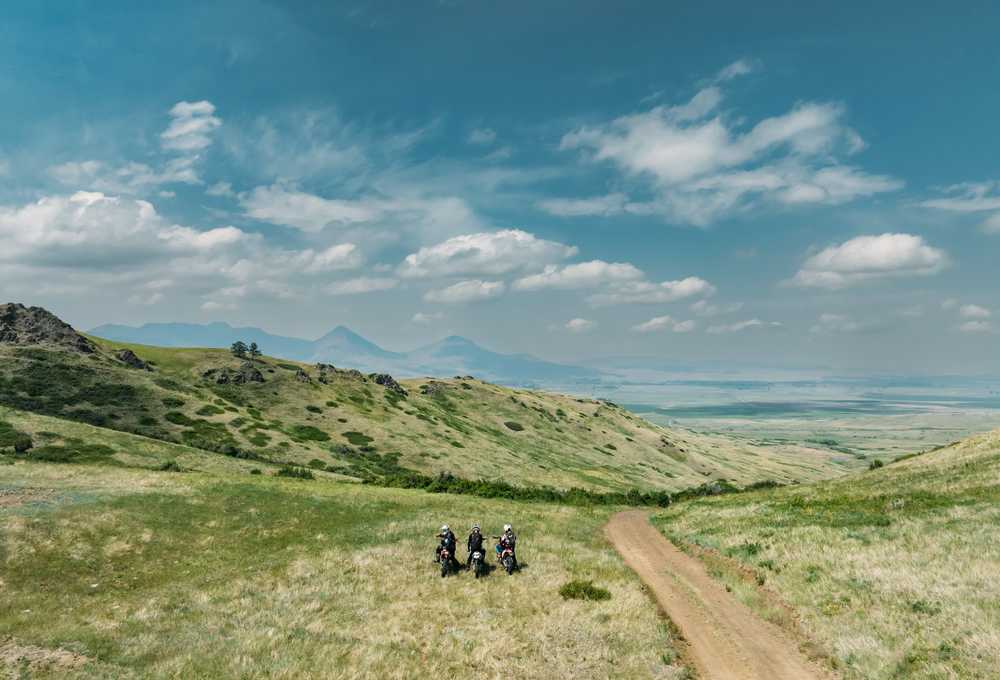 Small group hiking on a dirt trail through rolling green hills with distant mountains and a wide valley in Montana