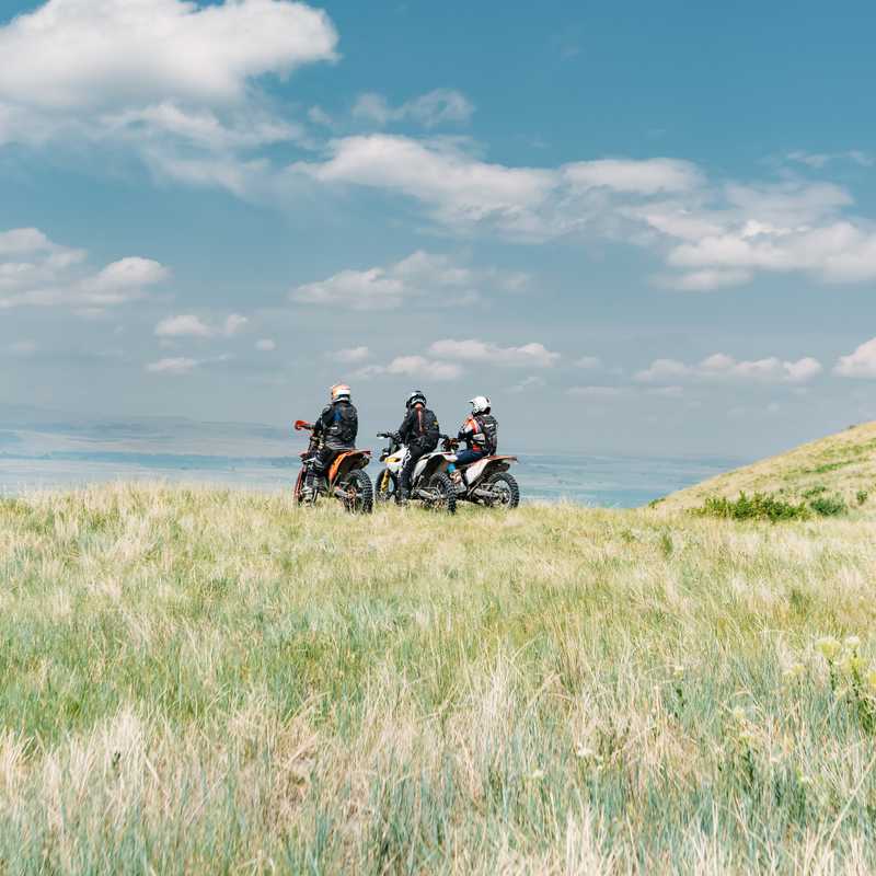 Motorcyclists on ATVs ride across a grassy hillside near the shoreline with mountains in the distance