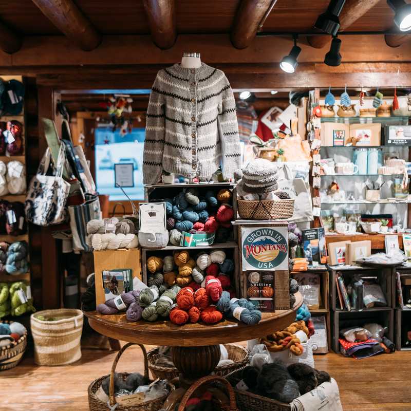 Gift shop display with folded sweaters, knit hats, yarn, and Montana “Grown in Montana” merchandise on shelves and a round table, with hanging shop lights above
