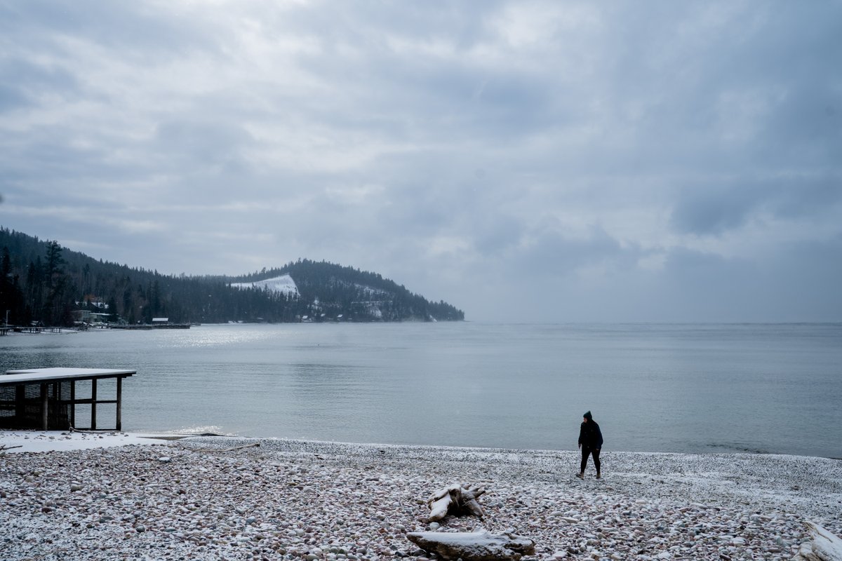 Person walking on a snowy shoreline along a calm lake in Montana with distant pine-covered hills and cloudy sky