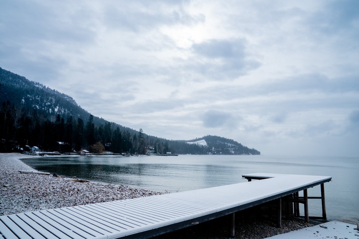 Snow-covered lakefront with a wooden dock extending into calm water under cloudy skies in Montana