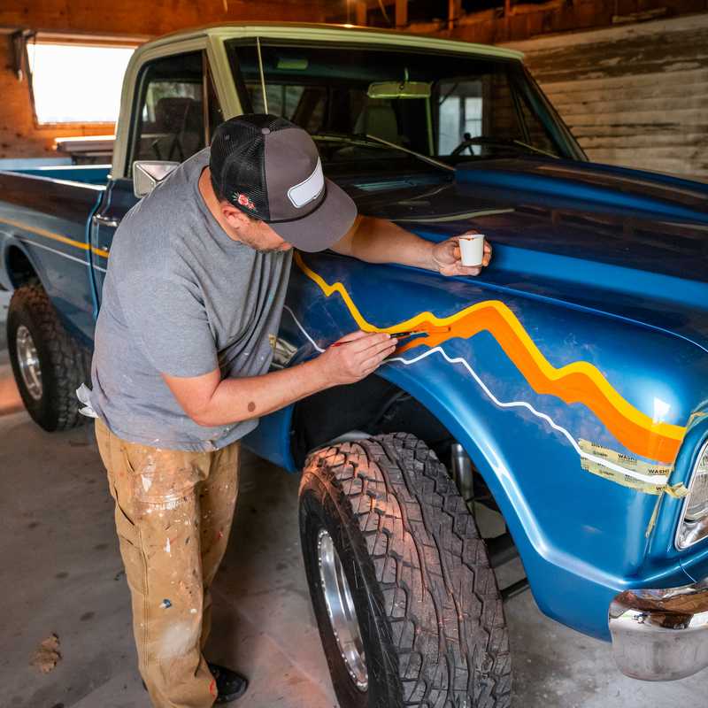 Man applying vehicle paint or graphics to a blue pickup truck in a garage with the hood open and tires visible