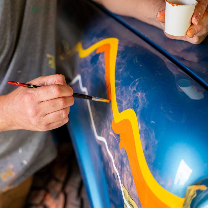 Person painting a blue car panel with a paintbrush and holding a cup while working outdoors